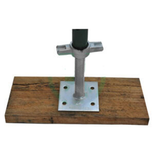 Scaffolding sole board with a base jack inserted through its center, sitting on a wooden plank. The base jack, with an adjustable threaded rod and a wing nut for height adjustment, is secured to the sole board, which distributes the load of the scaffolding to prevent sinking or damage to the ground surface beneath, ensuring stability and safety.